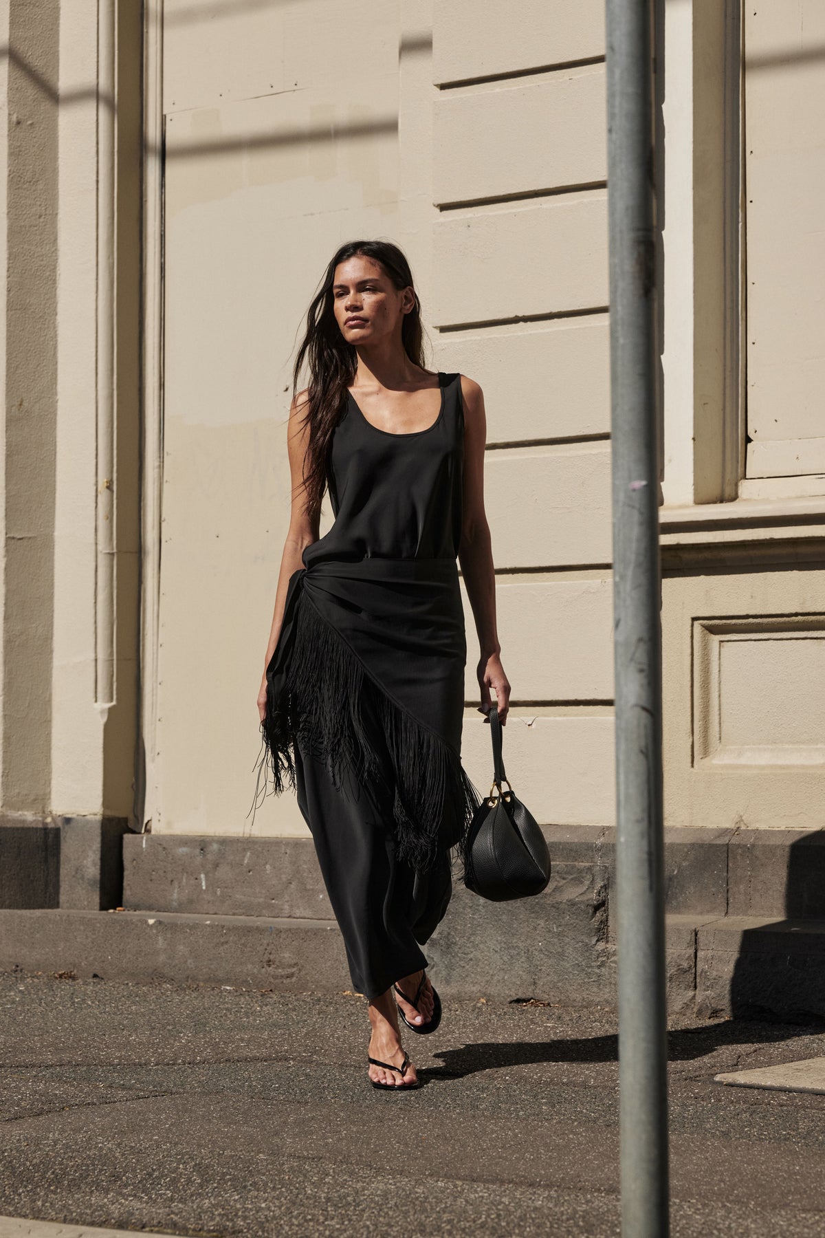 Woman wearing a black sleeveless maxi dress with fringe waist detail and carrying a handbag