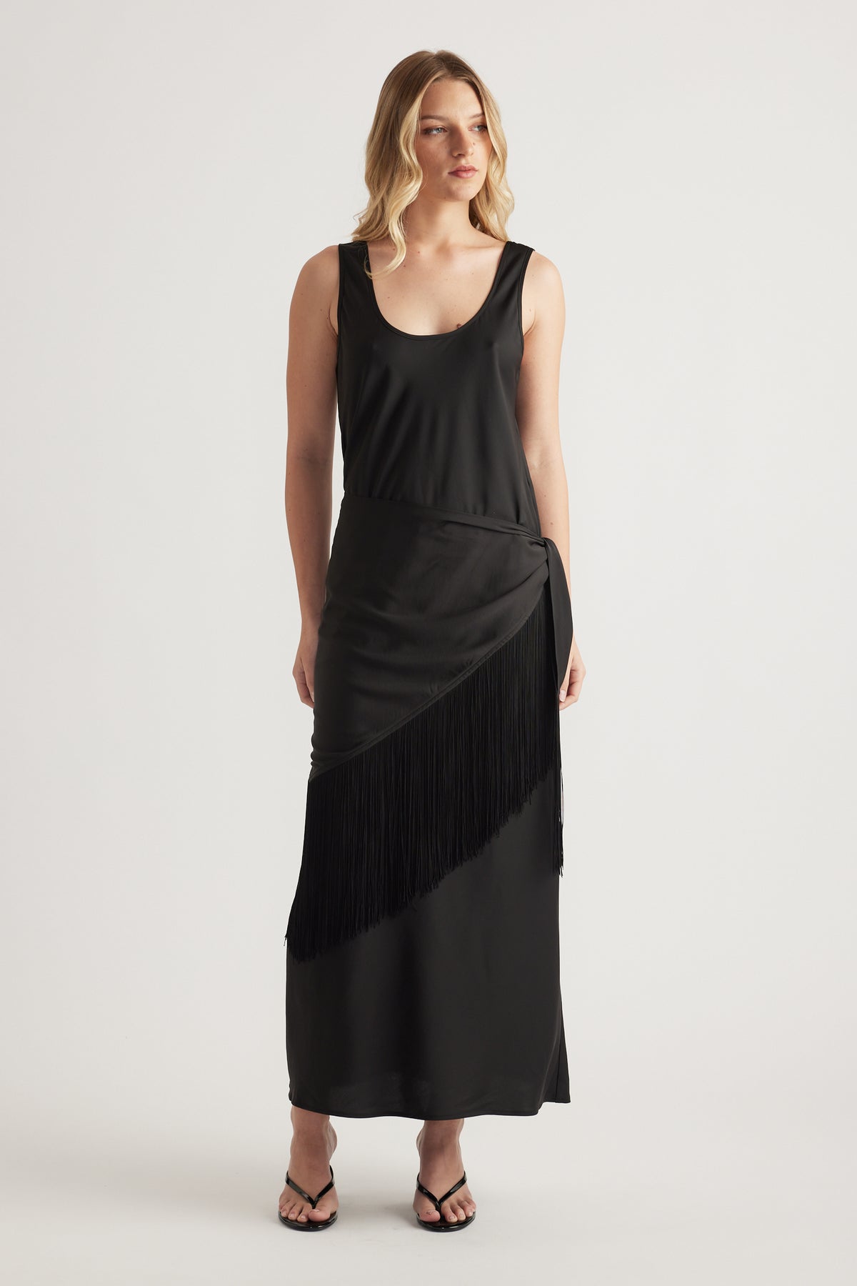 Woman wearing a black sleeveless maxi dress with a fringe waist and strappy sandals