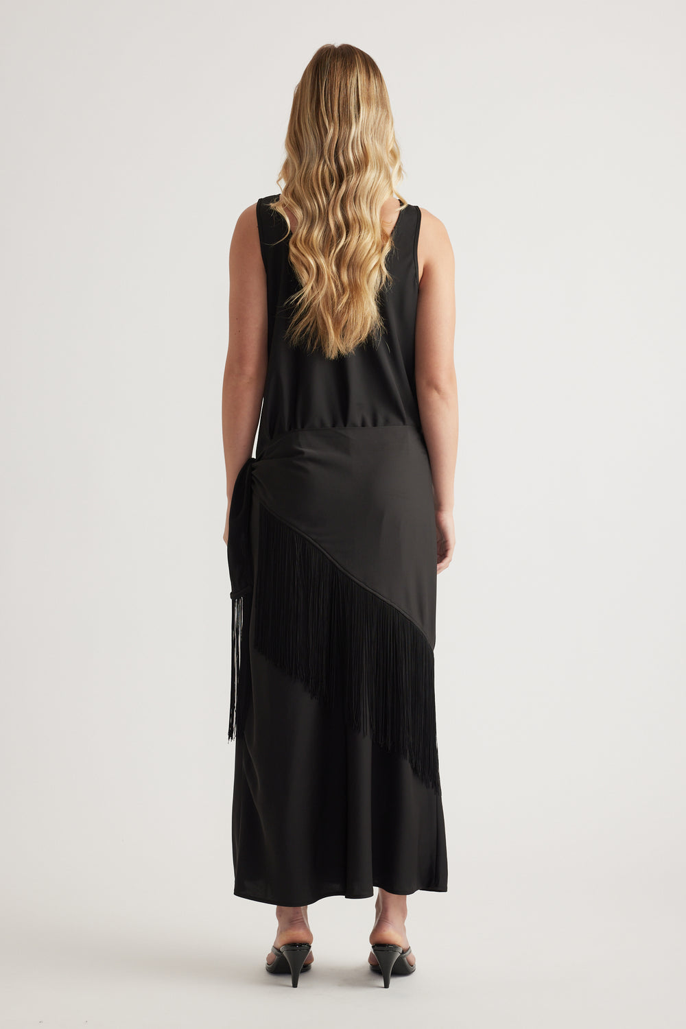 Back view of a woman wearing a black sleeveless maxi dress with a fringe waist scarf and kitten heels