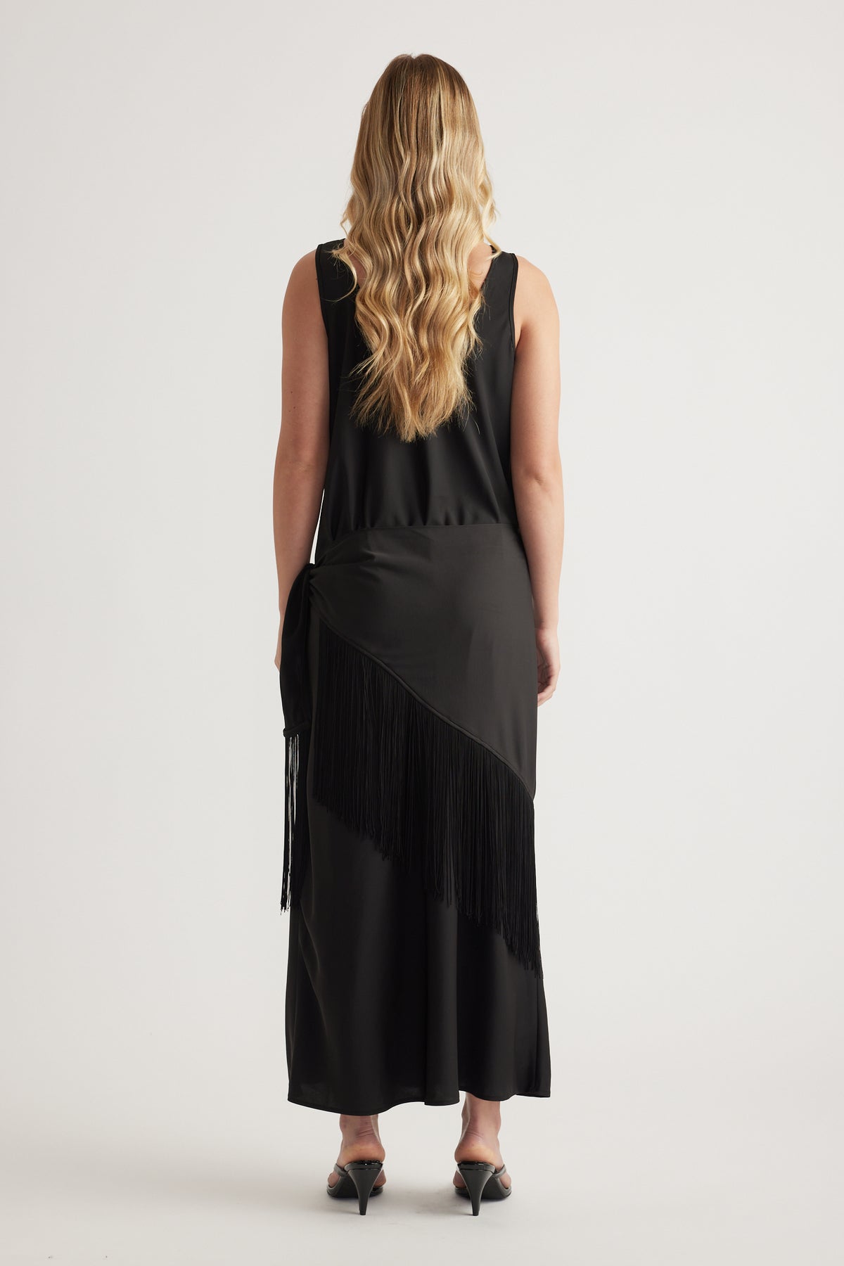 Back view of a woman wearing a black sleeveless maxi dress with a fringe waist scarf and kitten heels