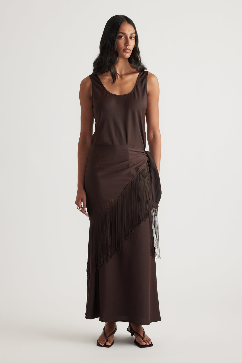 Woman wearing a dark brown sleeveless maxi dress with a fringe waist scarf and strappy sandals