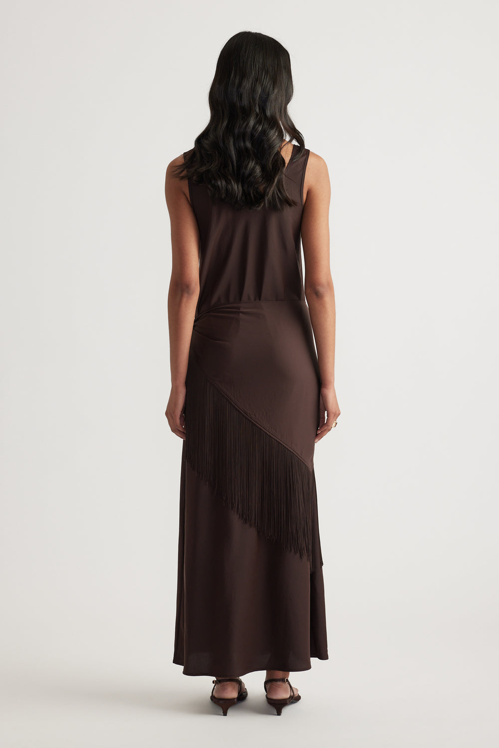 Back view of a woman in a dark brown sleeveless maxi dress with a draped fringe detail and black heels