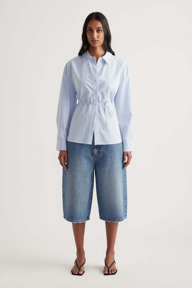 Woman wearing a blue and white striped collared shirt with waist tie, denim shorts and thong sandals