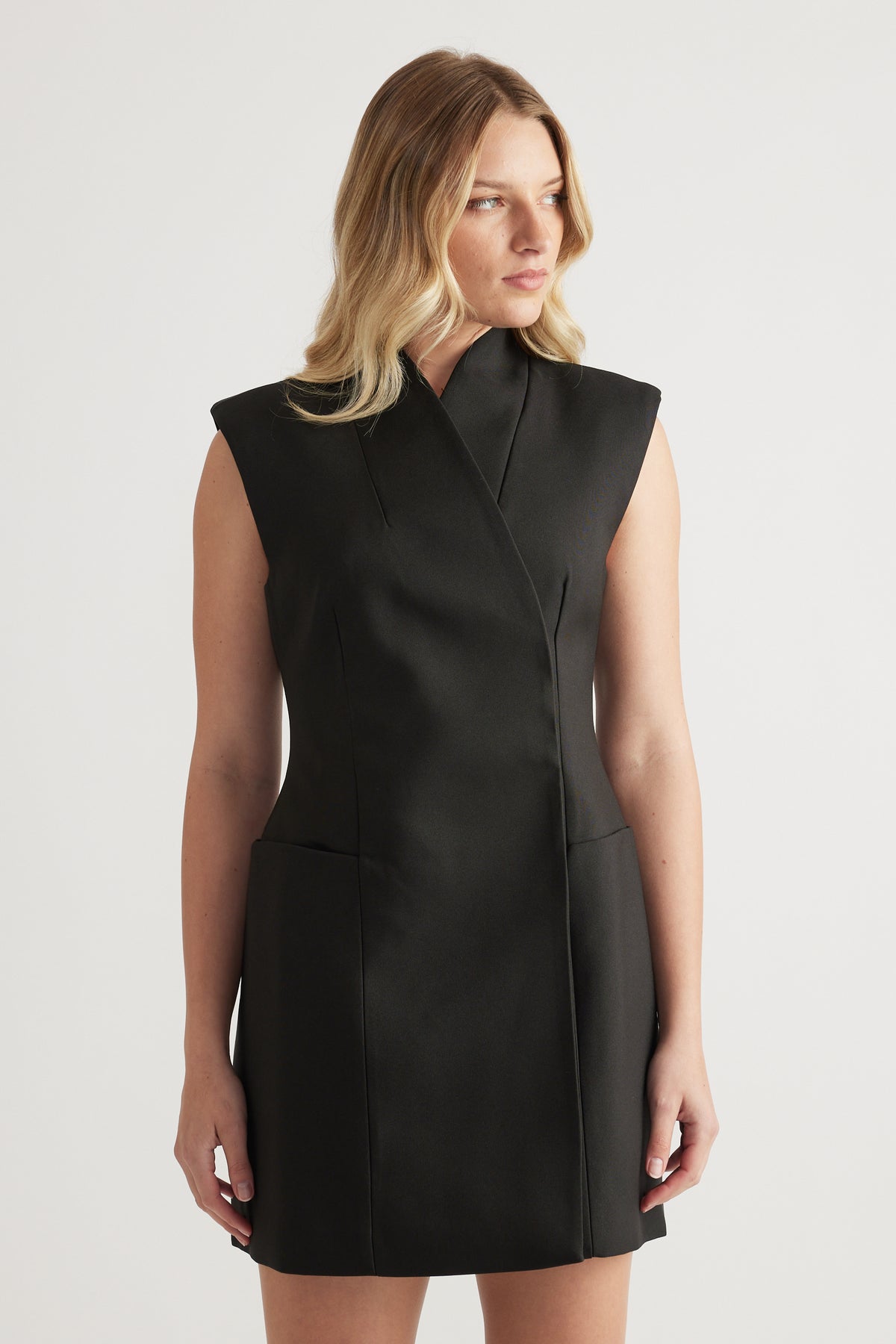 Woman wearing a black sleeveless high neck mini dress with structured paneling and front pockets