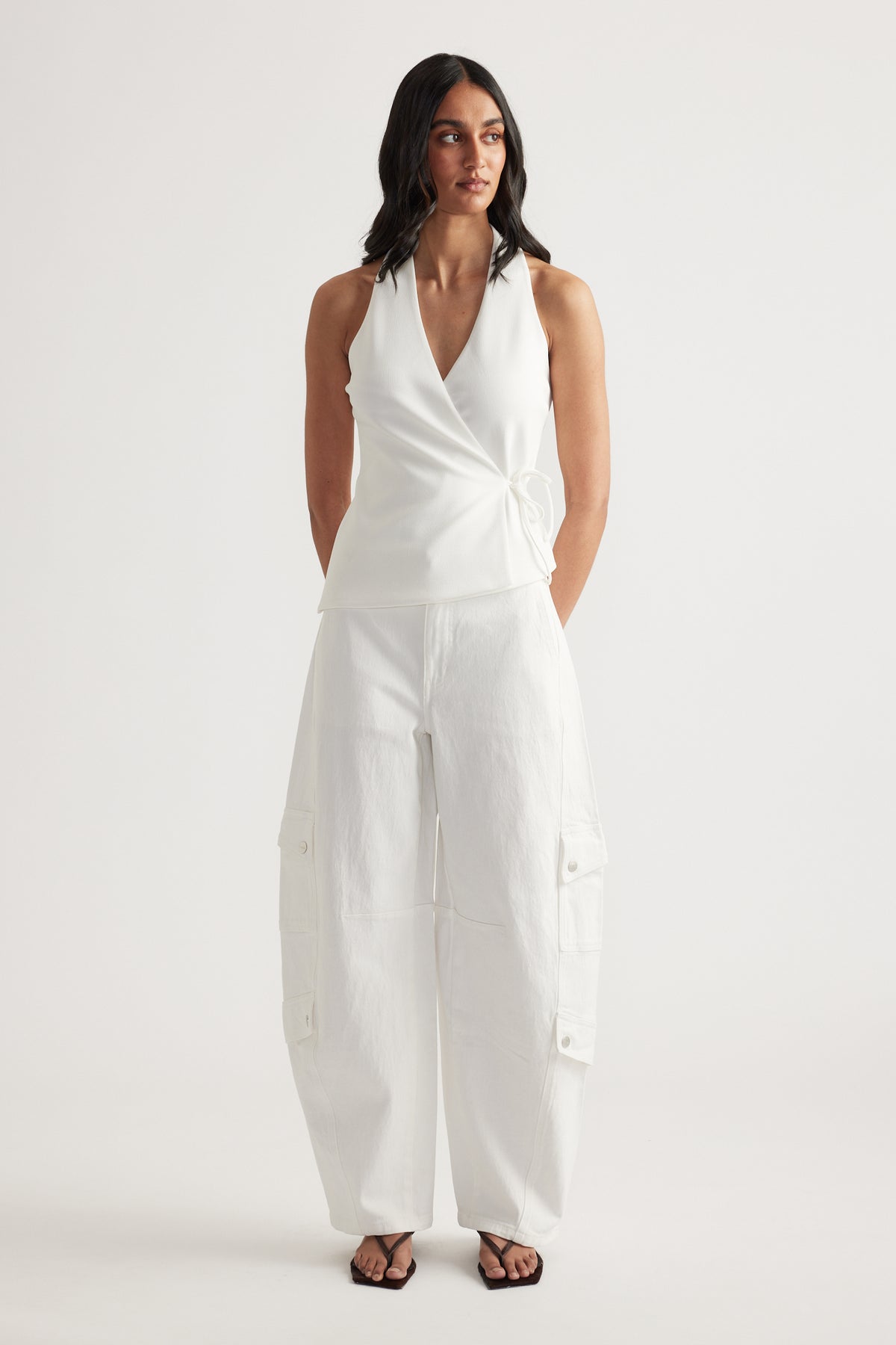 Woman wearing a white sleeveless wrap top with white cargo barrel leg jeans and black sandals