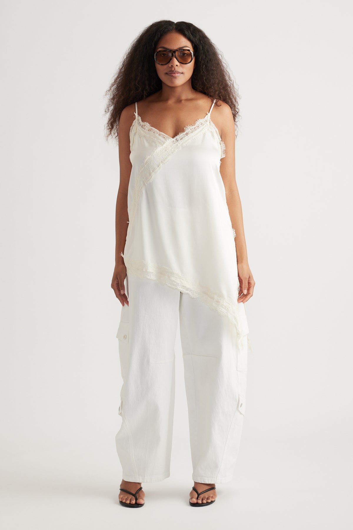 Woman wearing a white lace-trim camisole with relaxed cargo pants, sunglasses and strappy sandals