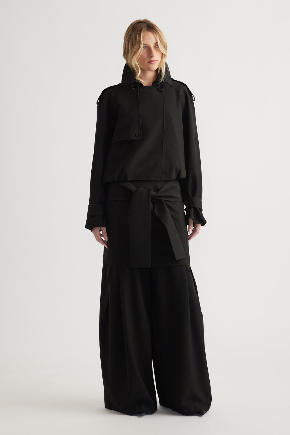 Woman wearing a black cropped bubble hem jacket layered over wide-leg black pants against a neutral background