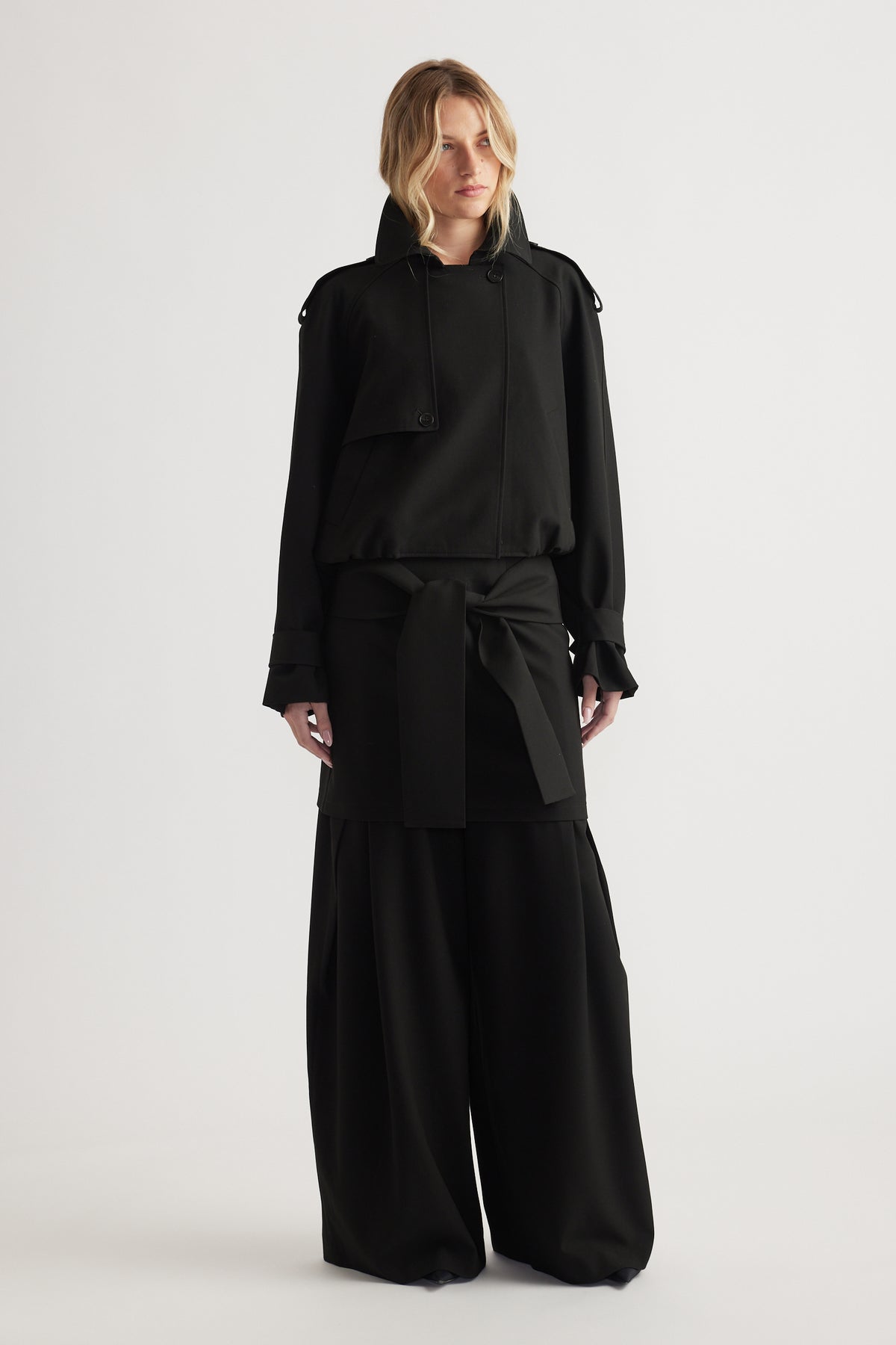 Woman wearing a black cropped bubble hem jacket layered over wide-leg black pants against a neutral background