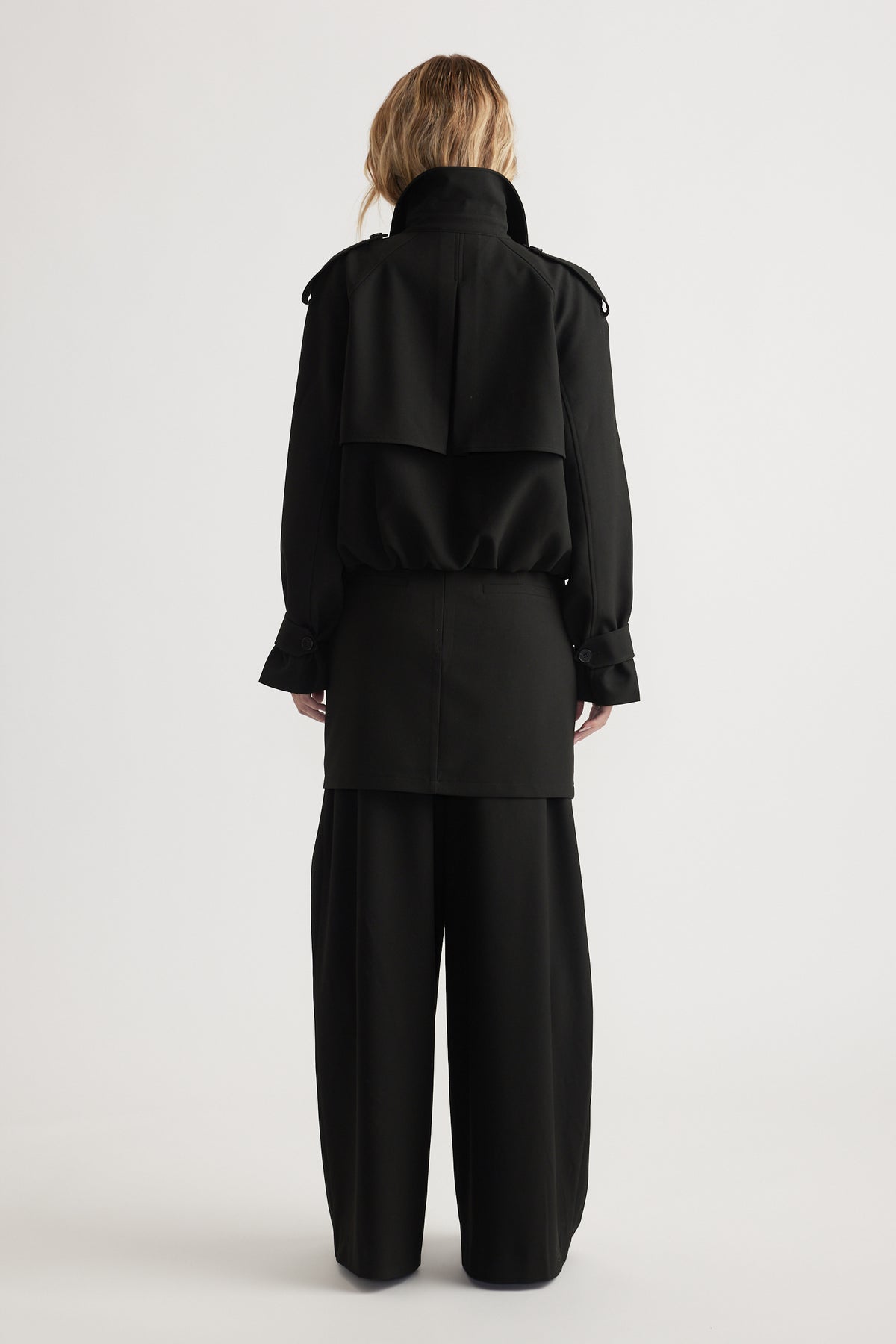 Back view of a woman wearing a cropped black trench coat with bubble hem and wide-leg black pants on a plain background