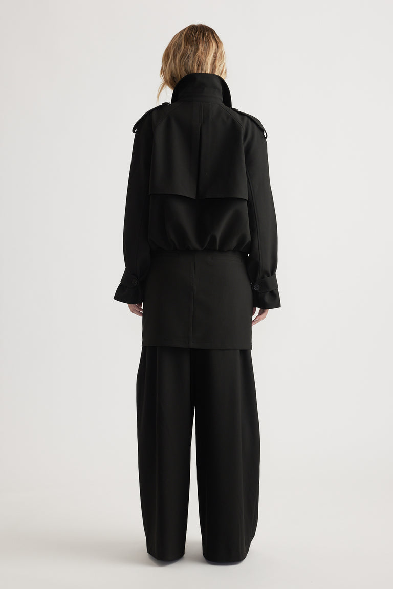 Back view of a woman wearing a cropped black trench coat with bubble hem and wide-leg black pants on a plain background