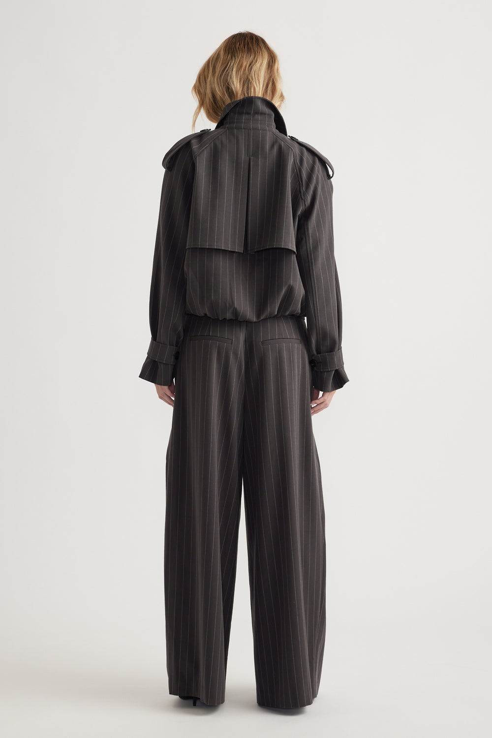 Rear view of woman wearing black pinstripe cropped trench with buttoned cuffs and matching wide-leg trousers