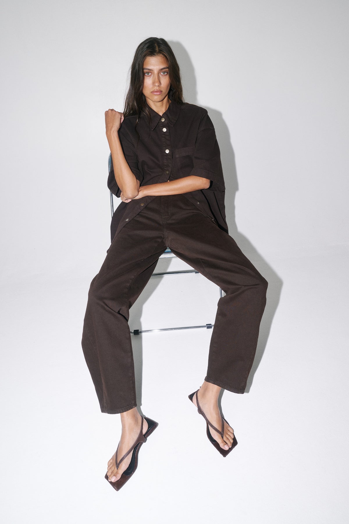 Woman seated in dark brown high-rise barrel-leg jeans with a matching shirt and thong sandals