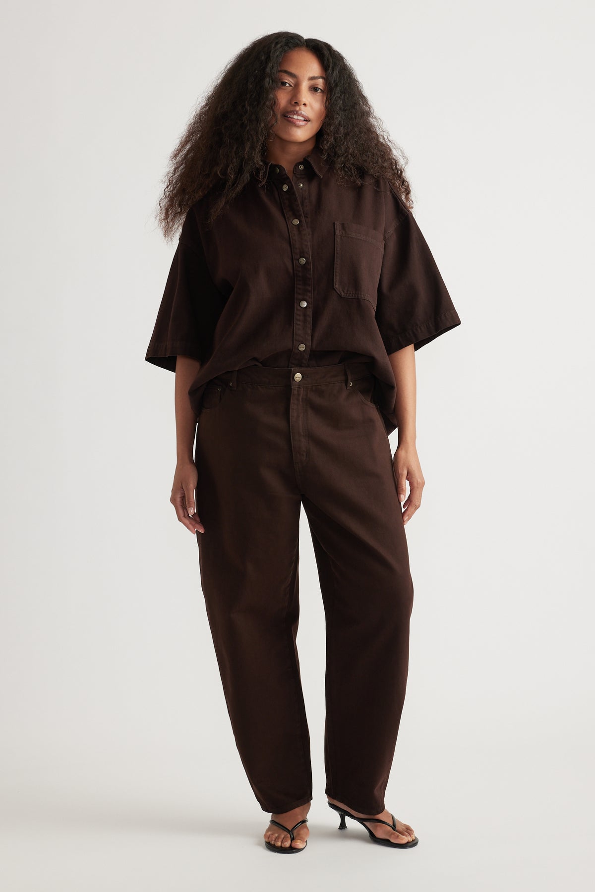 Woman wearing a dark brown oversized shirt with high rise barrel leg jeans and flat sandals