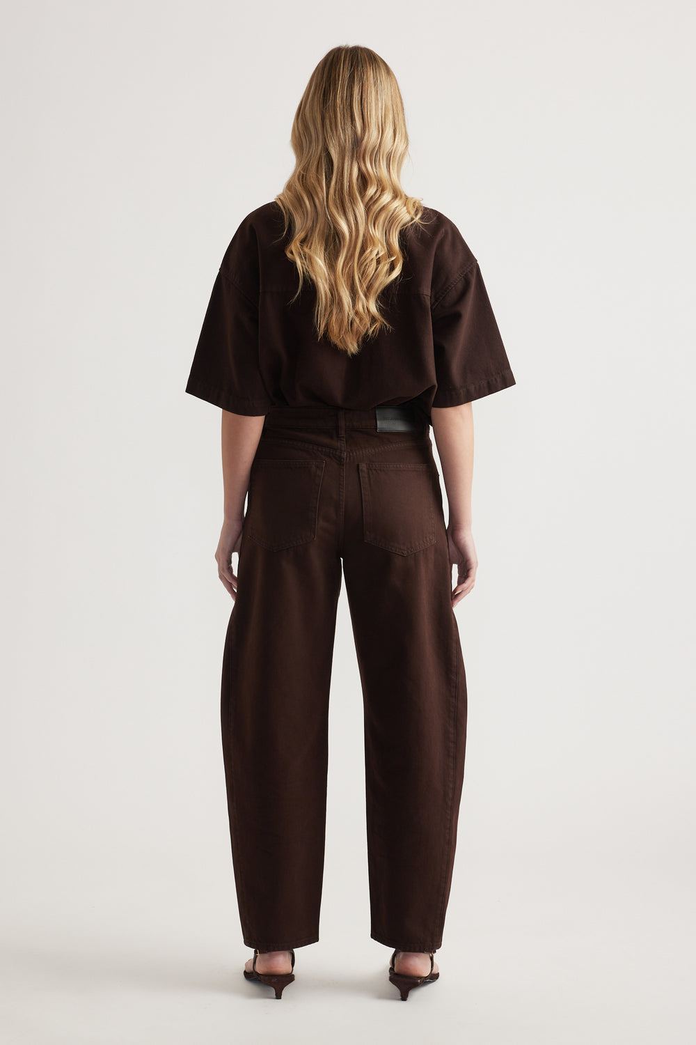 Back view of a woman wearing dark brown barrel leg jeans with a matching oversized tee and heels