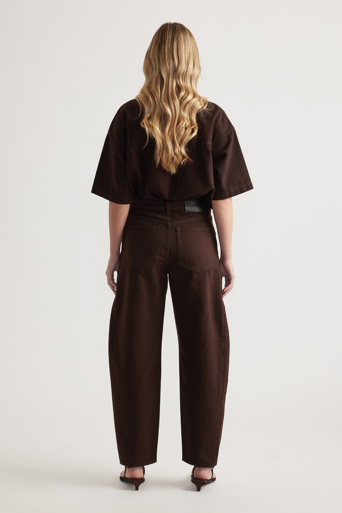 Back view of a woman wearing dark brown barrel leg jeans with a matching oversized tee and heels