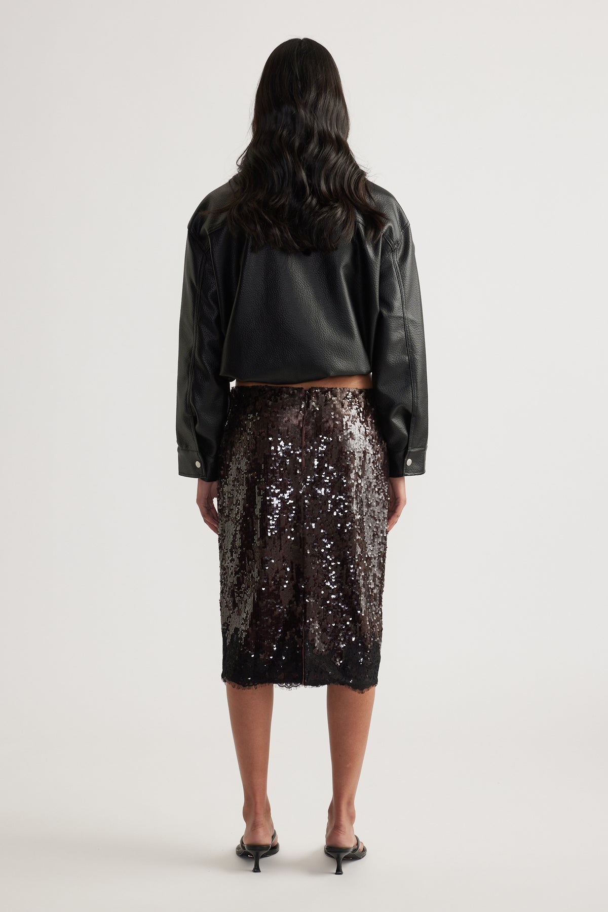 Woman viewed from behind wearing a black leather jacket, dark sequin knee length skirt and black heels