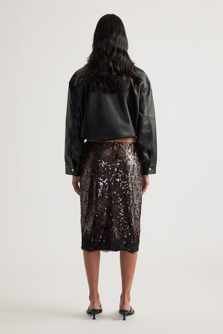 Woman viewed from behind wearing a black leather jacket, dark sequin knee length skirt and black heels