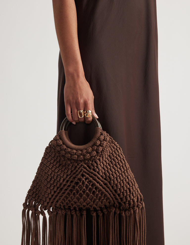 Close-up of a hand wearing layered stone rings and holding a brown macrame bag with fringe detailing