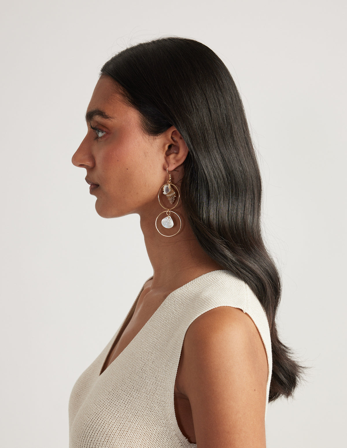 Woman wearing thin gold hoop earrings with speckled sea shell and mother of pearl details