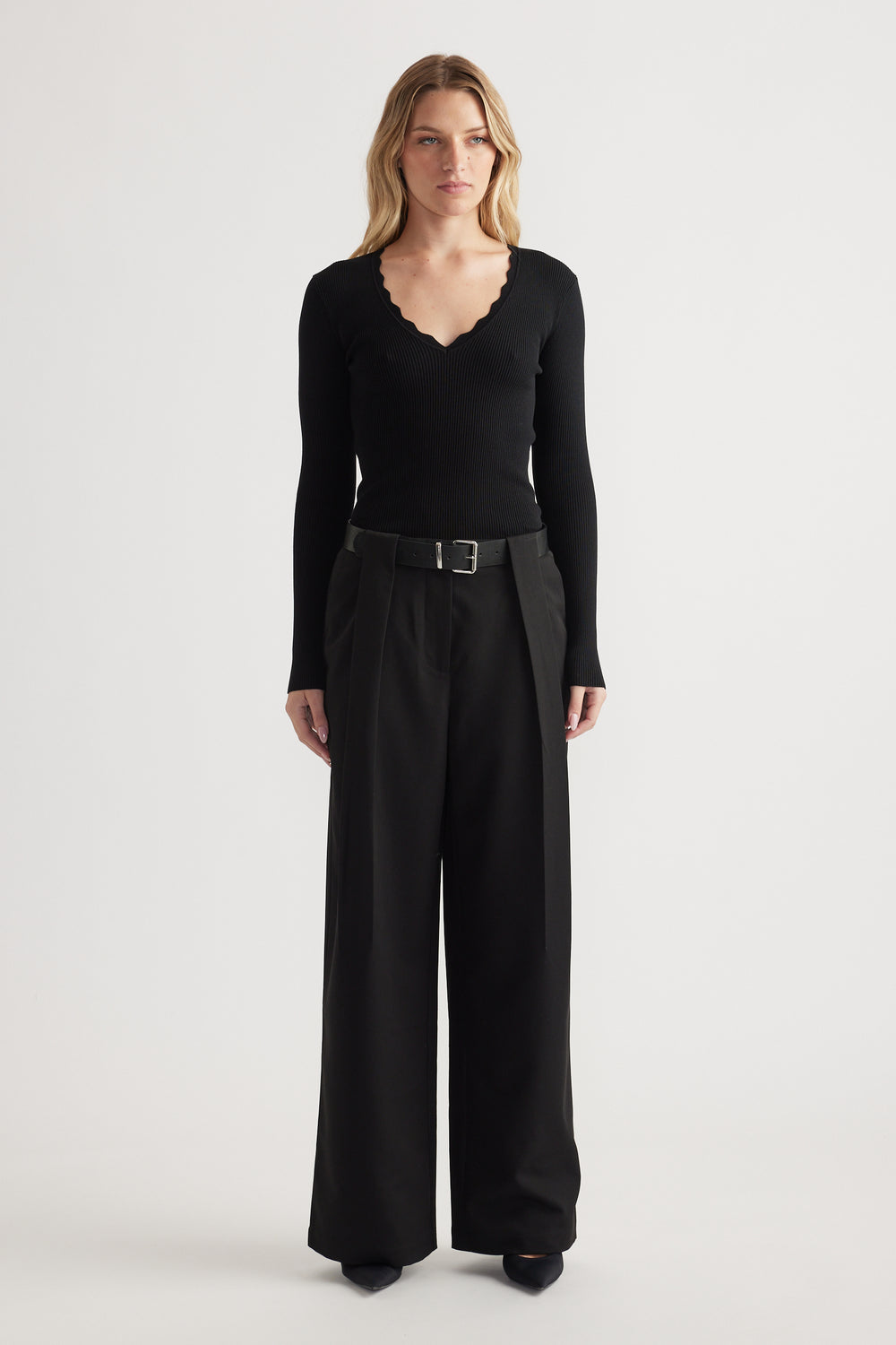 Woman wearing high-rise wide-leg black pleated trousers with a belt paired with a fitted scalloped-edge black top