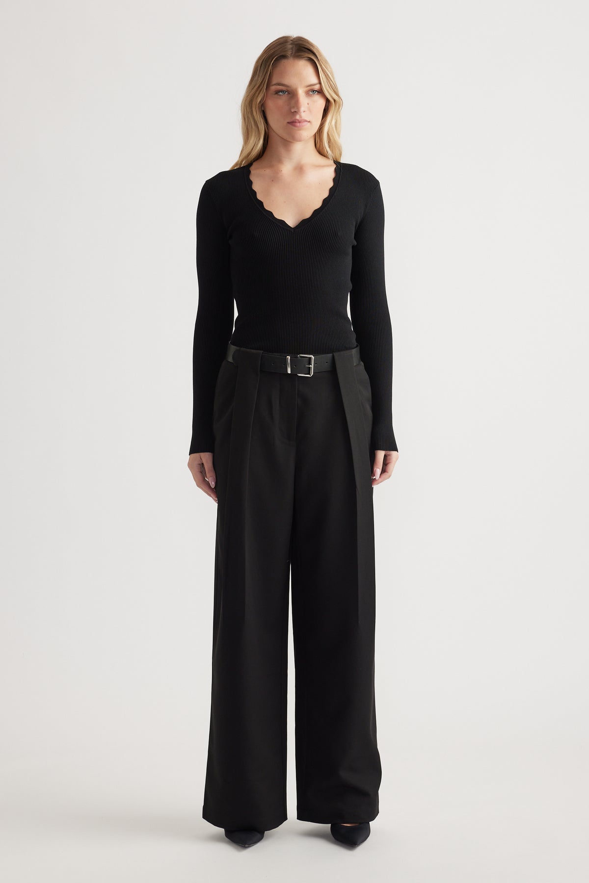 Woman wearing high-rise wide-leg black pleated trousers with a belt paired with a fitted scalloped-edge black top