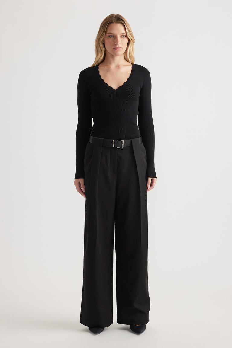 Woman wearing high-rise wide-leg black pleated trousers with a belt paired with a fitted scalloped-edge black top