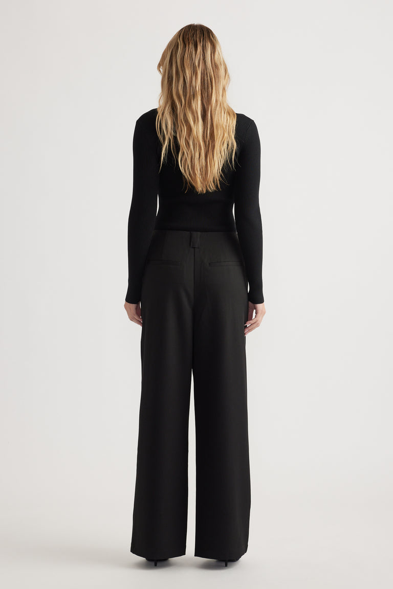 Woman with long wavy blonde hair wearing high-waisted black wide-leg pants and a fitted black long sleeve top, facing away