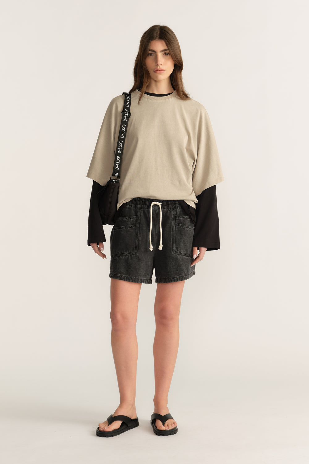 Woman wearing black denim shorts with a beige oversized tee layered over a black long sleeve top and sandals