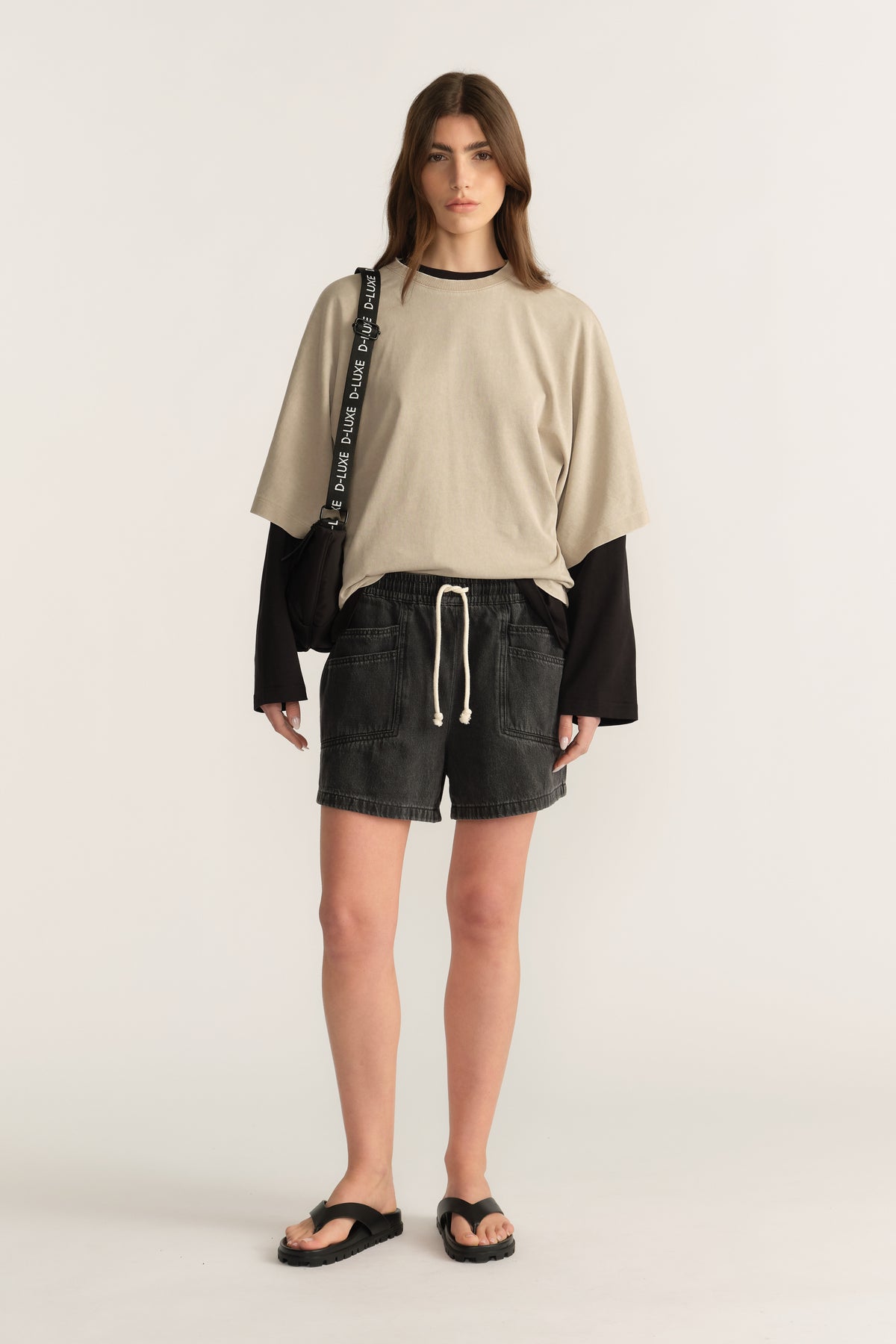 Woman wearing black denim shorts with a beige oversized tee layered over a black long sleeve top and sandals