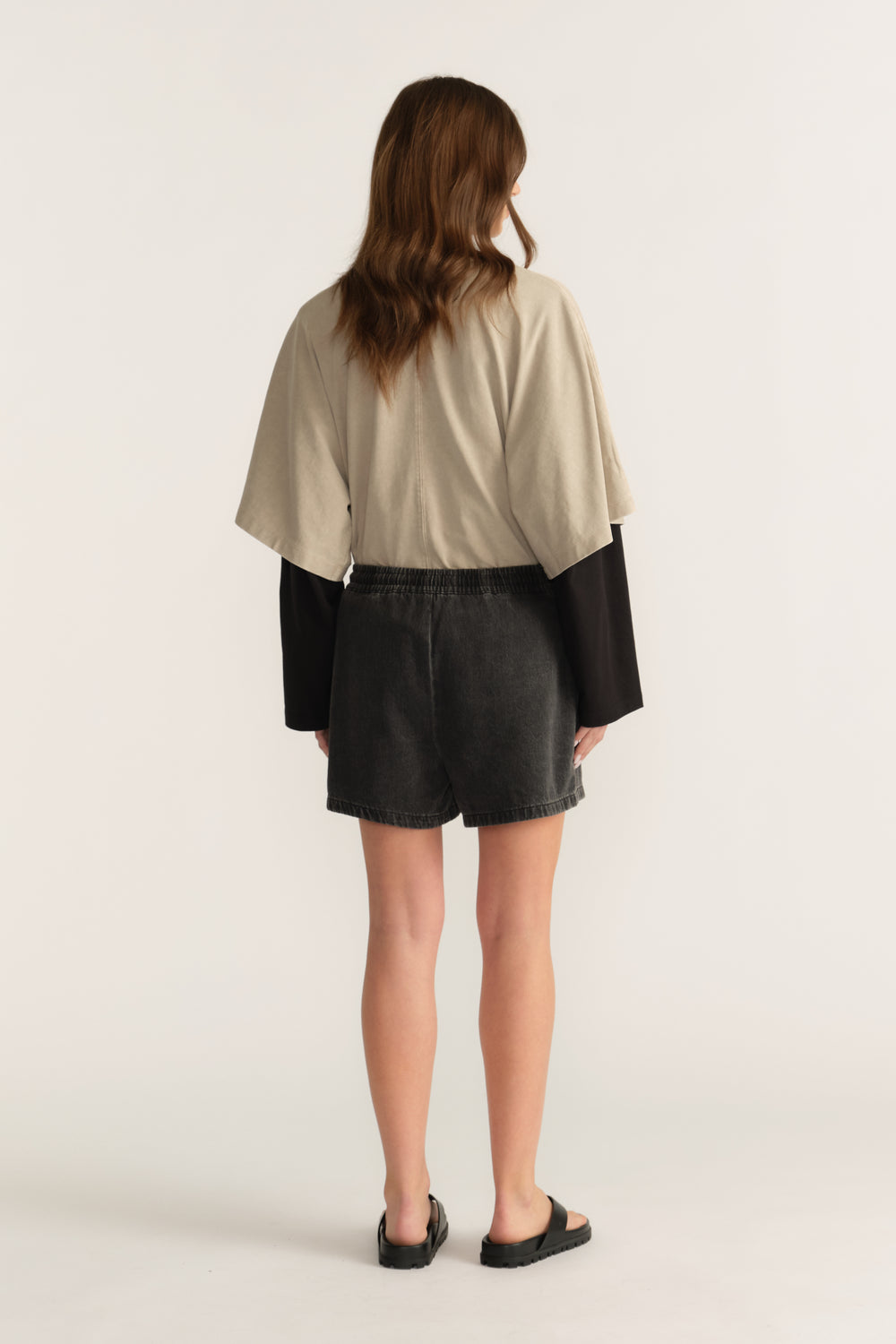 Back view of a woman wearing black denim shorts with a beige oversized tee layered over a black long sleeve top and sandals