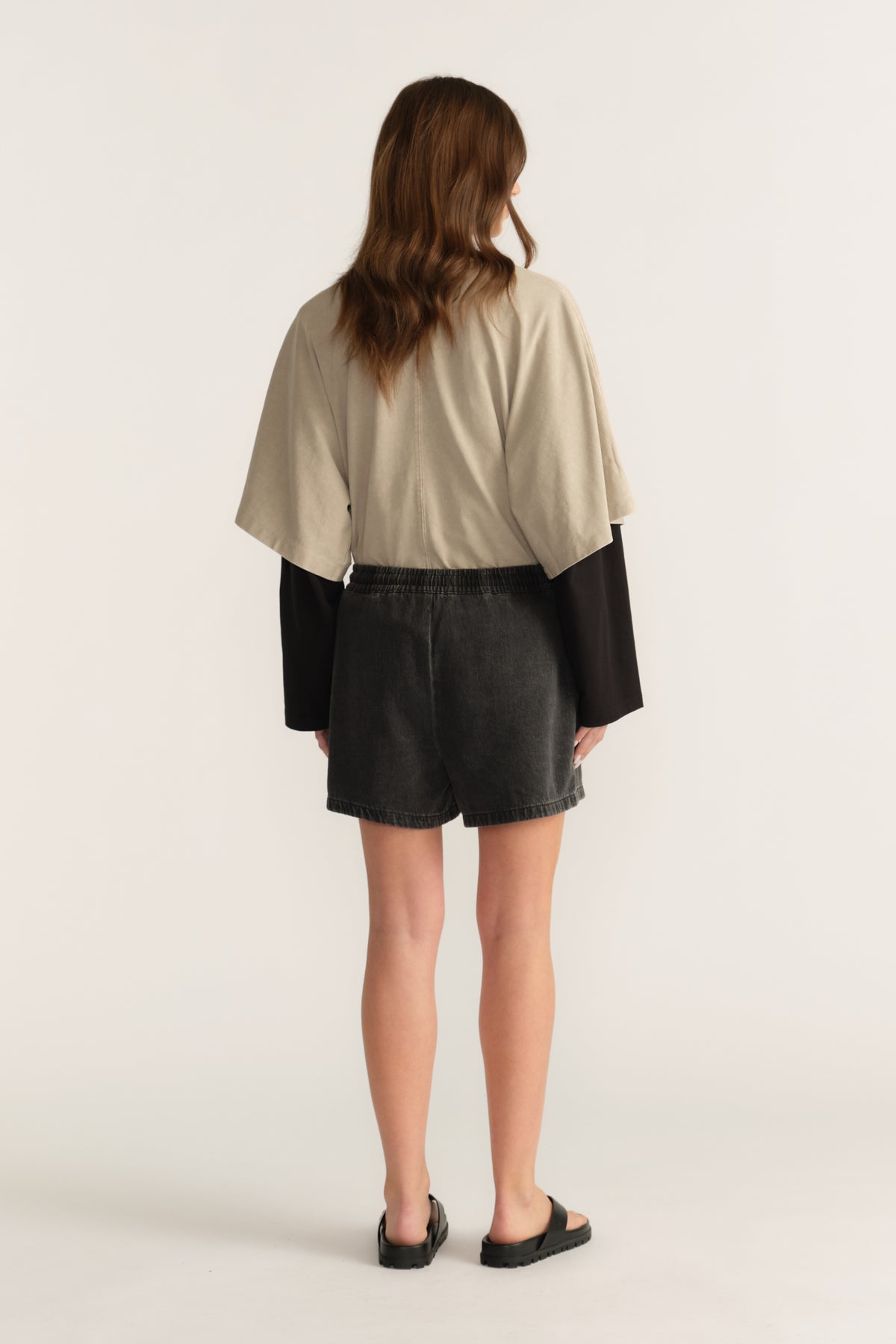 Back view of a woman wearing black denim shorts with a beige oversized tee layered over a black long sleeve top and sandals