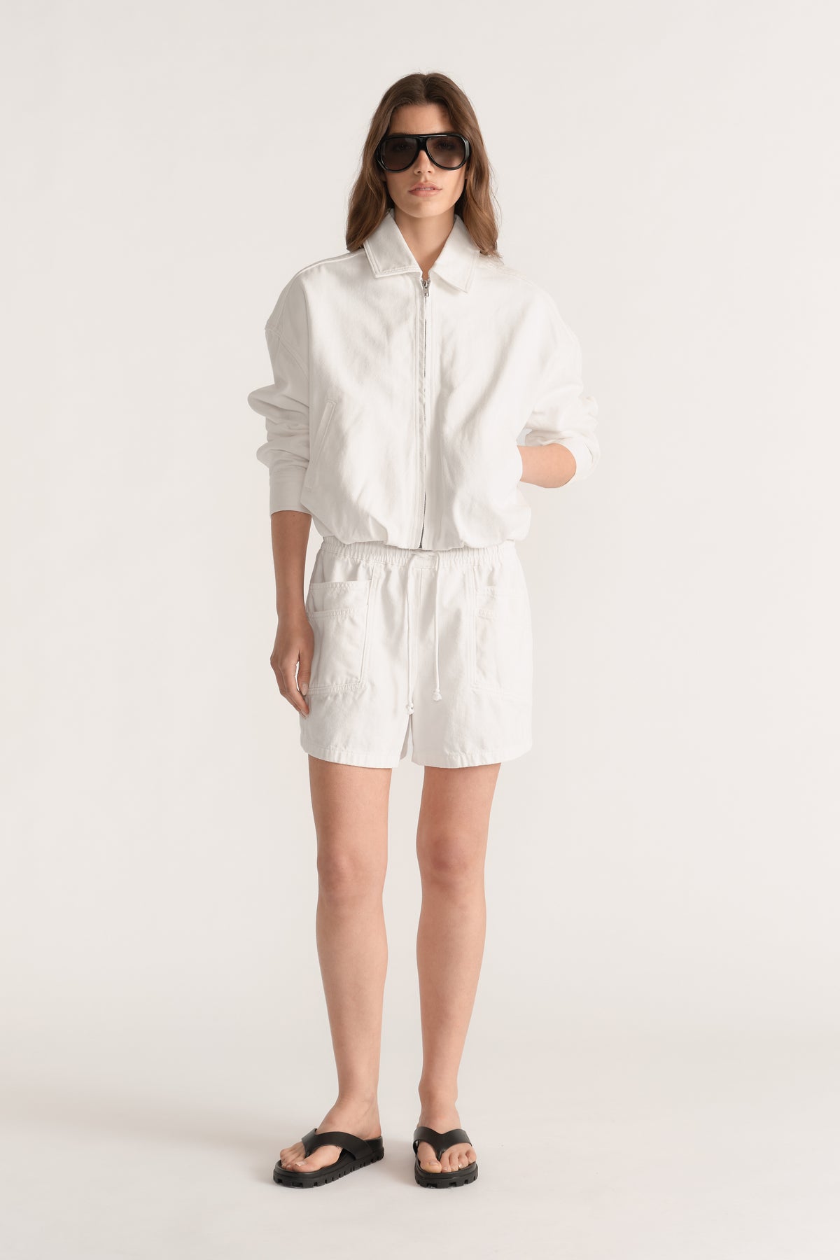 Woman wearing white denim pull on shorts with an elastic waist, zip jacket and black sandals