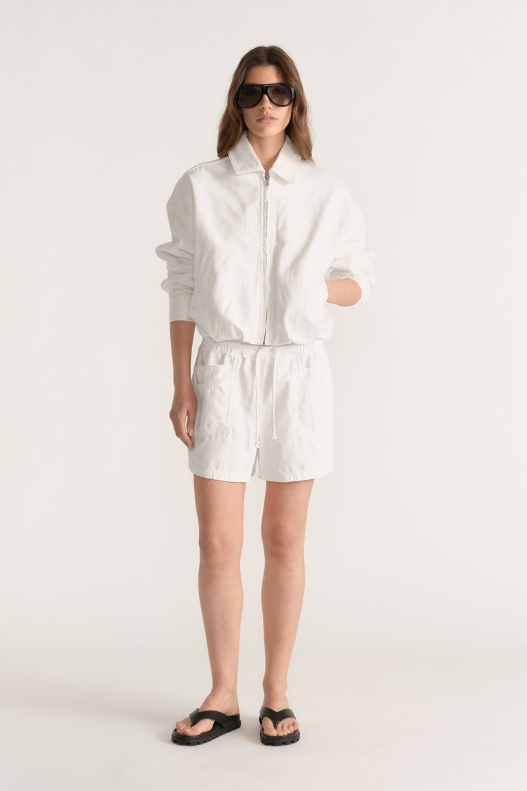 Woman wearing white denim pull on shorts with an elastic waist, zip jacket and black sandals