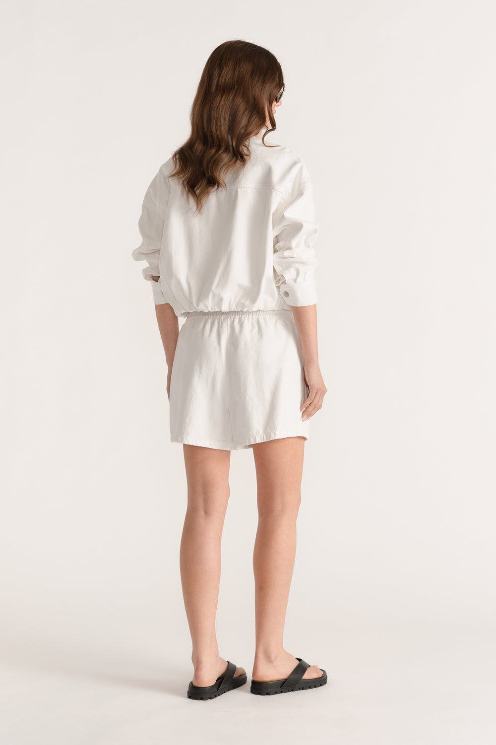 Model seen from behind wearing white elasticated denim shorts with a mid rise waist and black sandals
