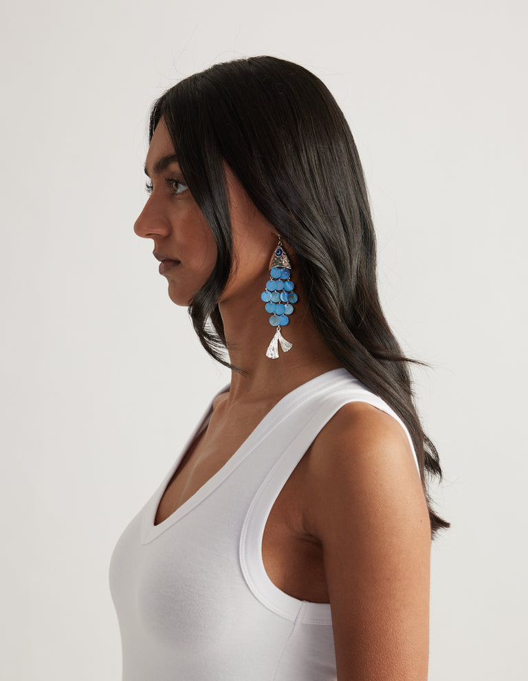 Woman in profile wearing blue pearlescent tiered fish earrings with silver detailing and a white tank top