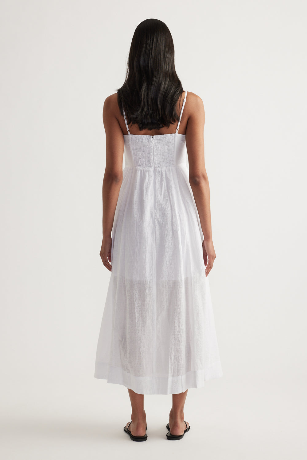 Woman viewed from behind wearing a white strappy midi dress with a cinched waist and flowing fit and flare silhouette