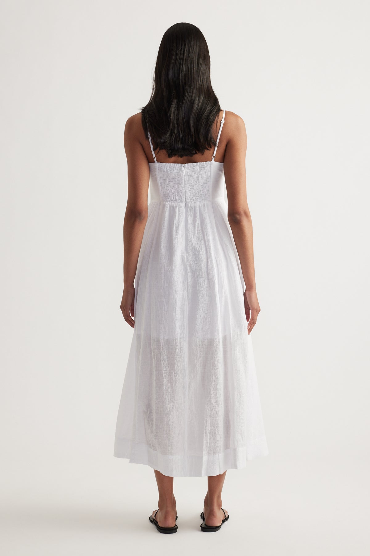 Woman viewed from behind wearing a white strappy midi dress with a cinched waist and flowing fit and flare silhouette