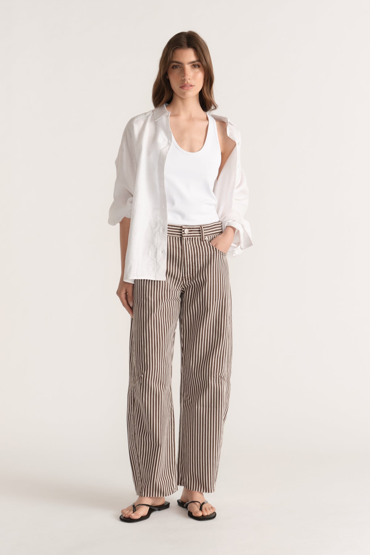 Woman wearing striped barrel-leg jeans with a white tank, oversized shirt and black flat sandals