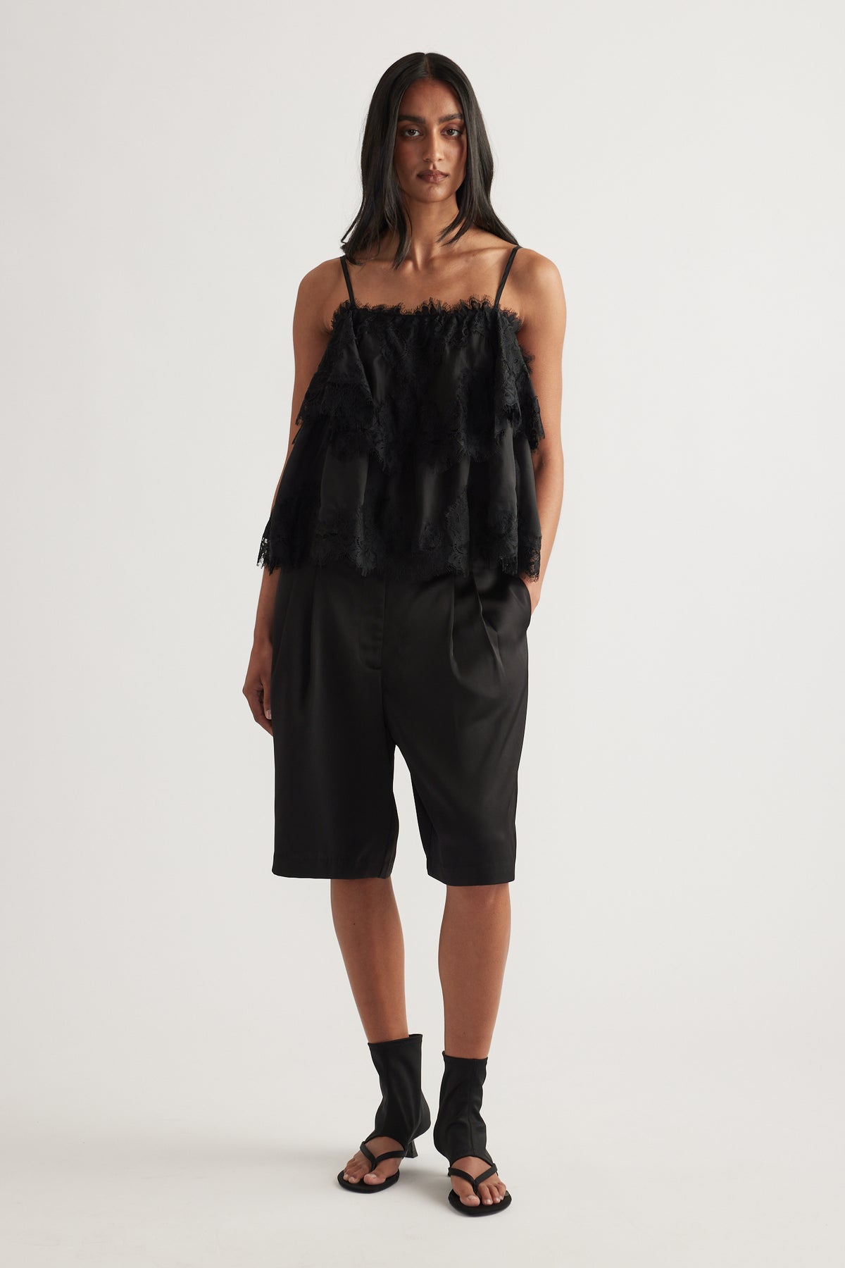 Woman wearing black tailored satin shorts with pleat detail, styled with a fringed camisole and sandals