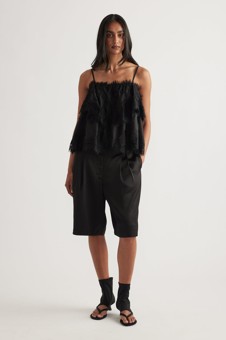 Woman wearing black tailored satin shorts with pleat detail, styled with a fringed camisole and sandals