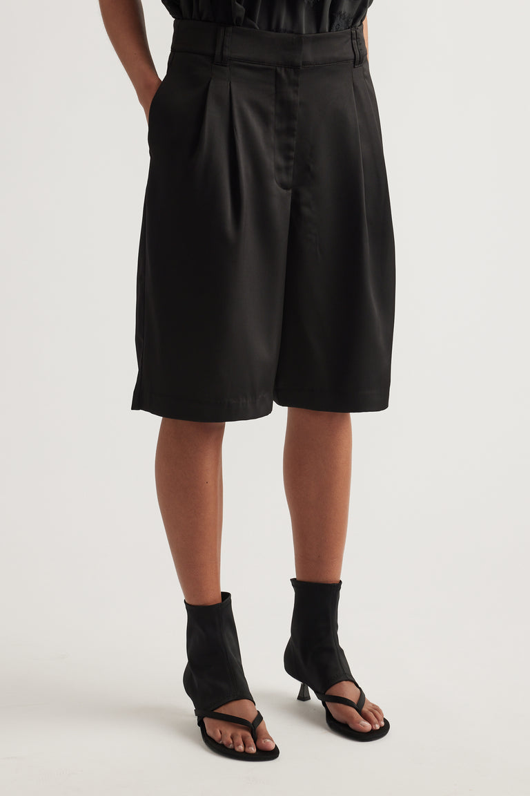 Woman wearing black satin tailored shorts with pleats and pockets, styled with a black top and heeled sandals