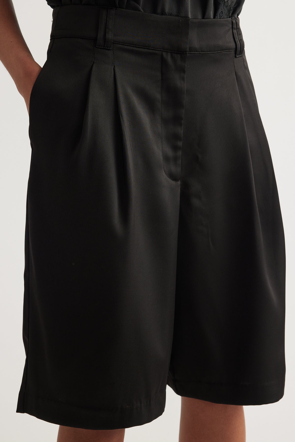 Woman wearing black satin tailored shorts with pleats, a mid-rise waist and above-knee hem