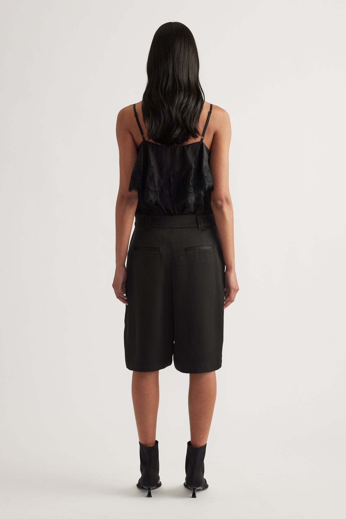 Woman viewed from behind wearing black tailored satin shorts with pleats and a black camisole
