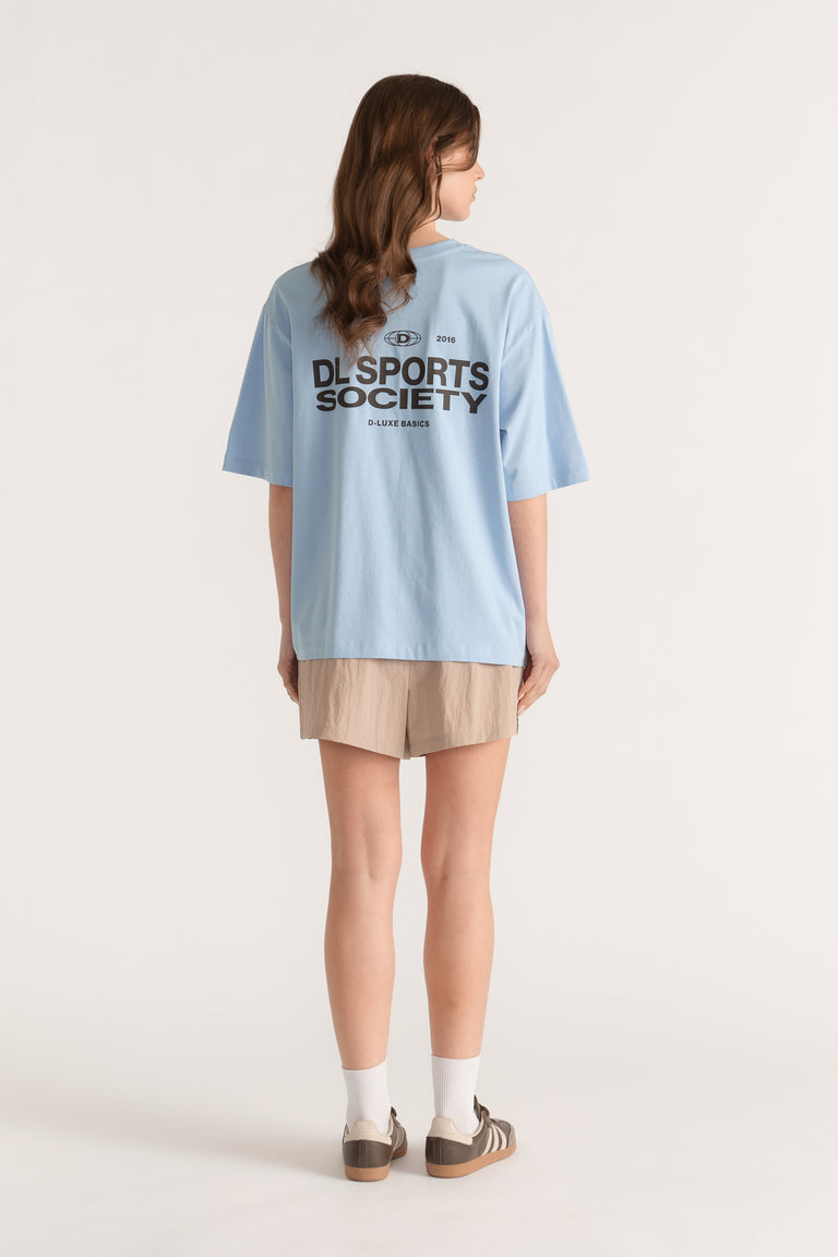 Back view of model wearing a light blue D Luxe sports graphic tee with beige shorts and sneakers