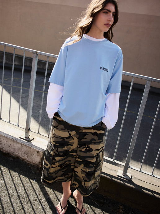 A woman stands outdoors near a metal railing, wearing an oversized light blue crewneck sweatshirt layered over a white long-sleeve shirt, paired with camouflage cargo shorts and black sandals.