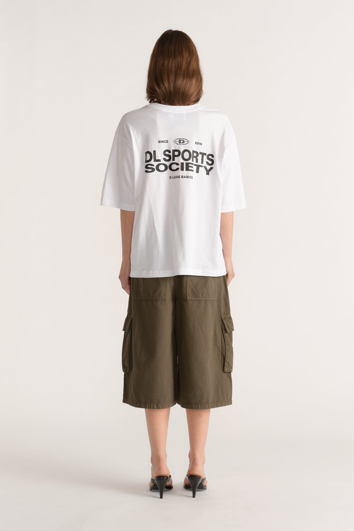 Model wearing a relaxed white graphic tee with olive cargo shorts and black heeled sandals viewed from behind