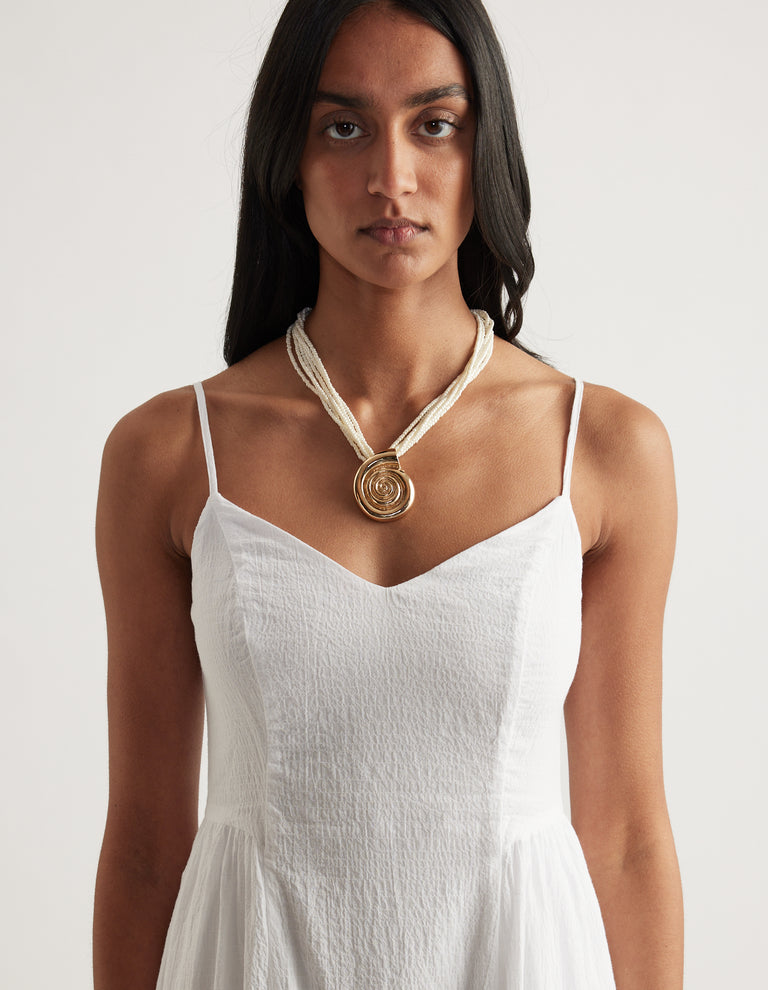 Woman wearing a pearlescent shell layered bead necklace with a circular pendant over a white strappy dress