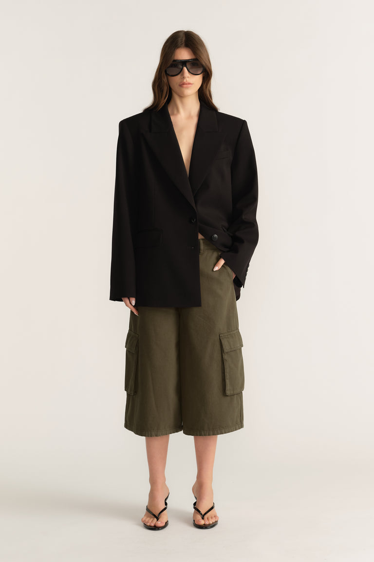 Woman wearing a black blazer with olive cargo shorts, sunglasses and thong sandals