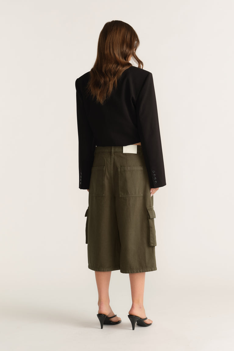 Woman wearing olive cargo shorts with side pockets, a black blazer and heeled sandals, viewed from behind