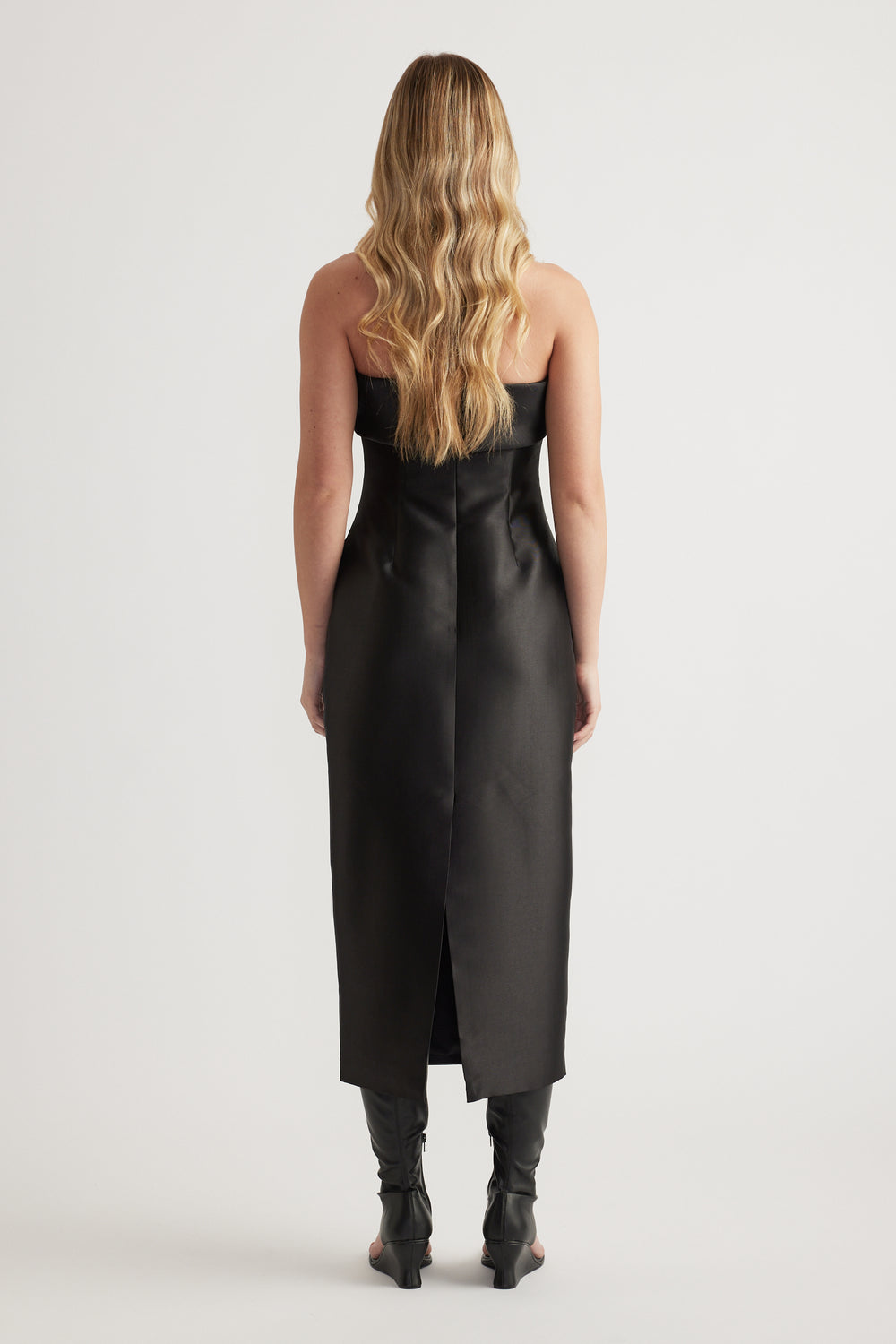 Woman wearing a fitted black midi dress with a straight neckline and ankle boots, viewed from behind